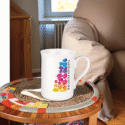 Weiße Keramiktasse mit farbenfrohem Würfeldesign und vertikalem Schriftzug auf einem runden Mosaiktisch vor einem Sofa – anschauliches Beispiel für bedruckte Tassen Werbemittel