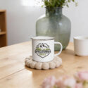 Emaillebecher mit silbernem Rand und Berg-Logo auf Holztisch mit Vase und Blumen
